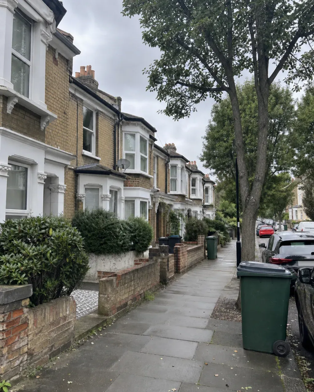 A quiet Balham street of yellow stock brick Victorian terraces with white bay windows, clipped hedges behind low brick walls and wheelie bins on a damp pavement