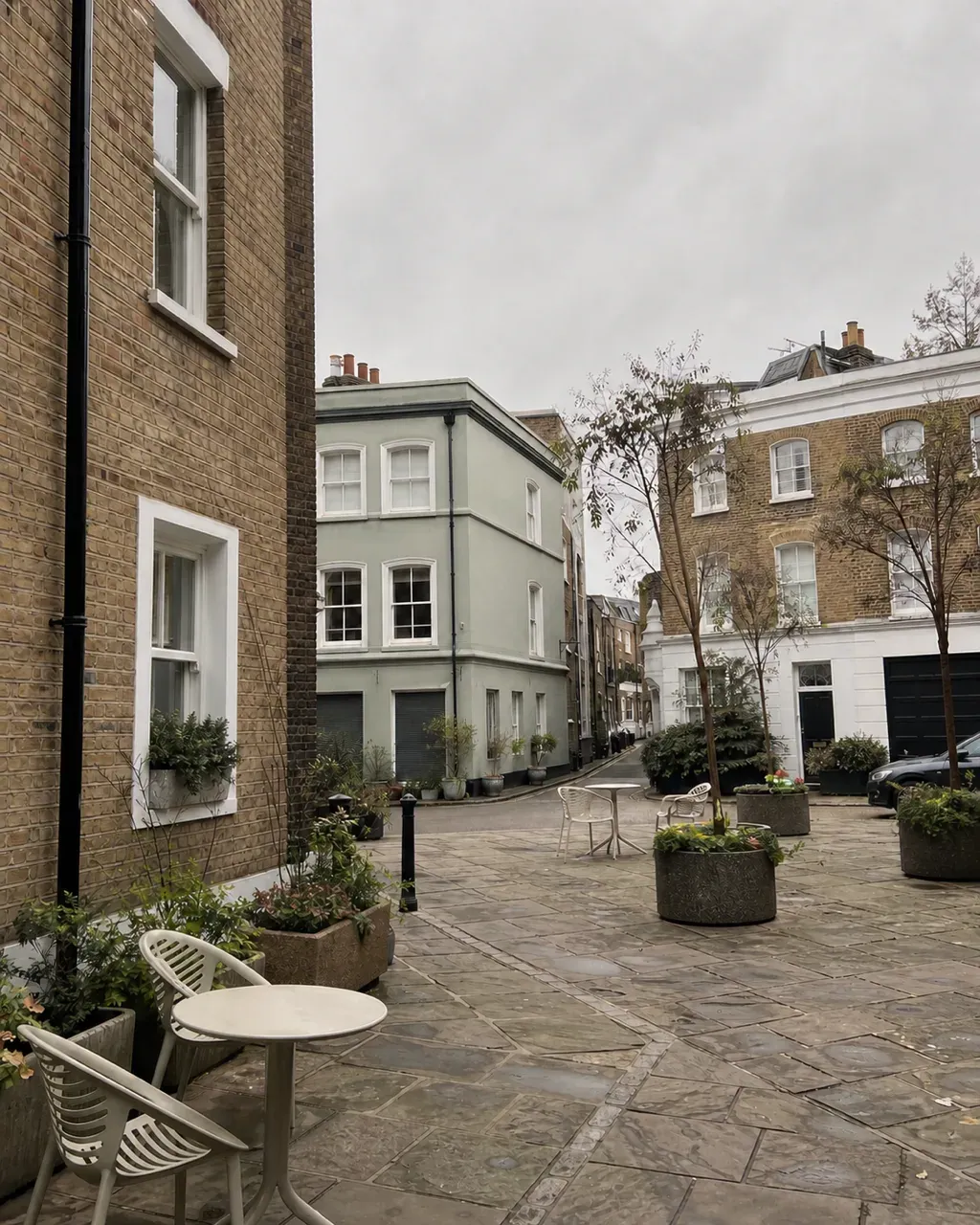 A paved corner at Battersea Square enclosed by low brick and painted stucco buildings, with stone planters, young trees and empty cafe tables on the flagstones