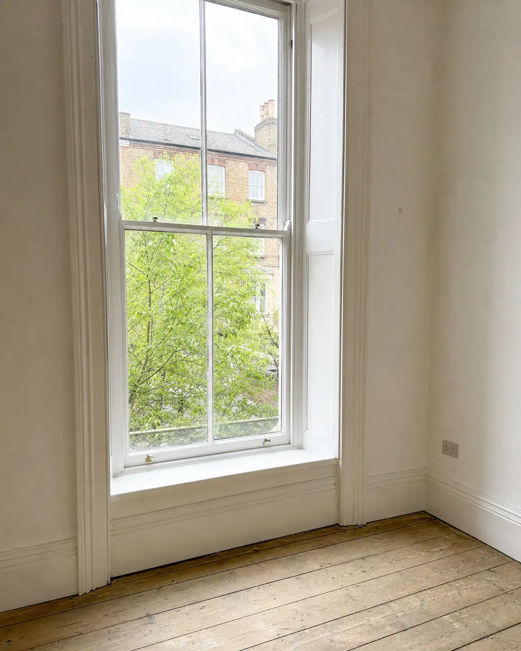 A tall sash window and clean bare sill in an empty Clapham conversion flat, with stripped floorboards and a London stock brick terrace visible through the glass