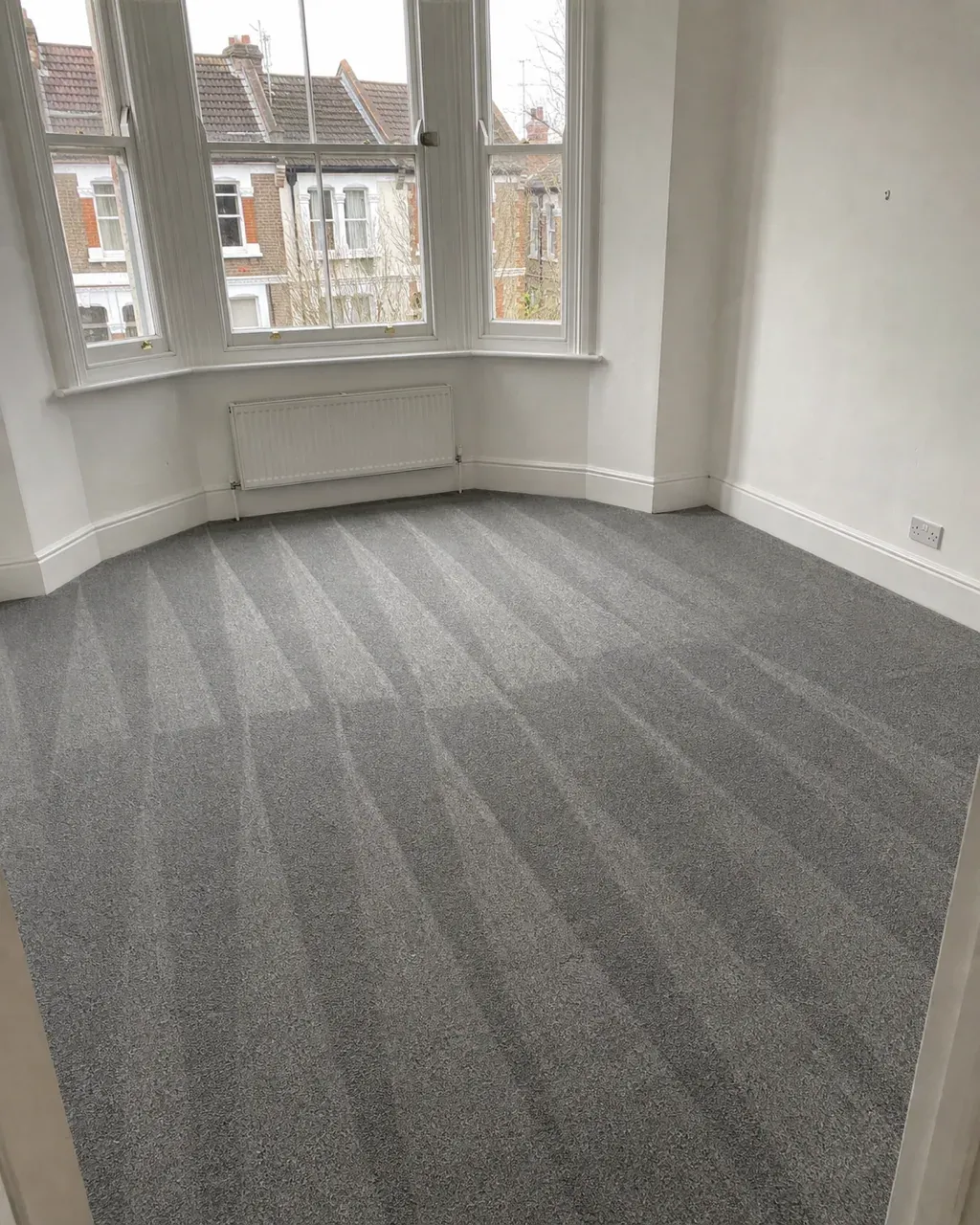 An empty Clapham living room with fresh hoover lines across grey carpet running towards a bay sash window with a radiator beneath it