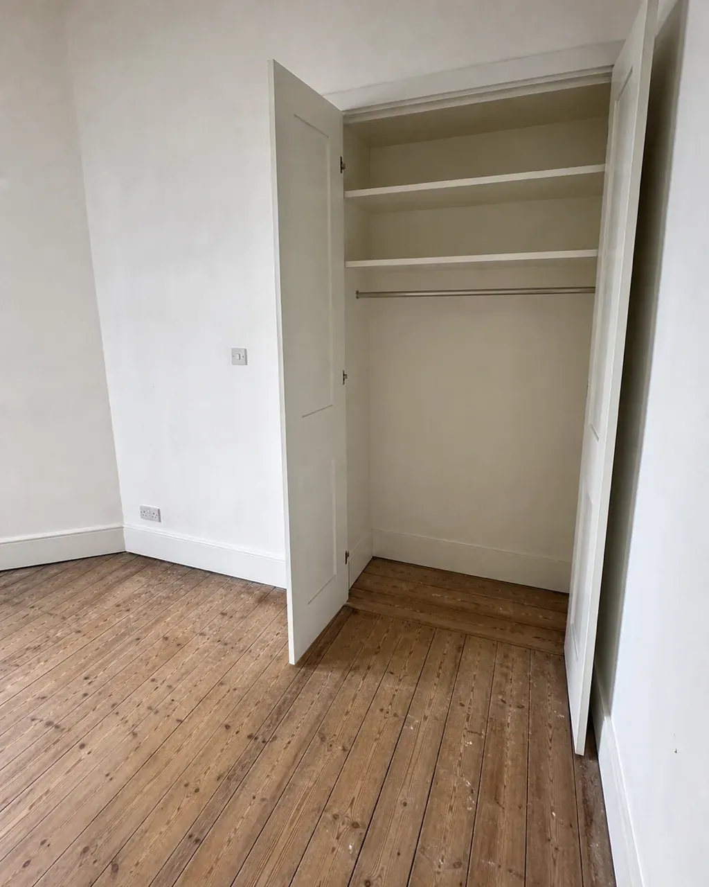 A fitted wardrobe standing open in an empty Clapham bedroom, its shelves and hanging rail completely bare and wiped clean above stripped floorboards